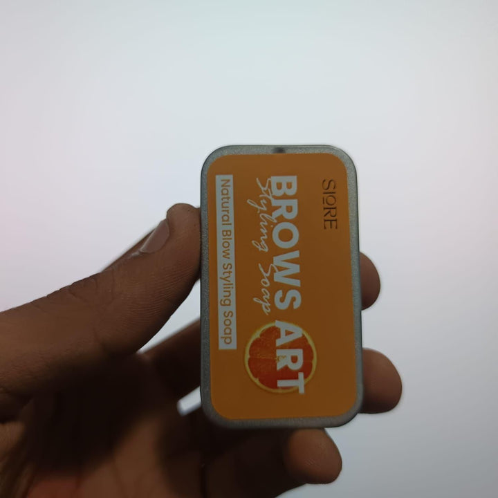 Hand holding SIORE BROWS ART Styling Solid Perfume natural brow styling soap in orange rectangular tin
