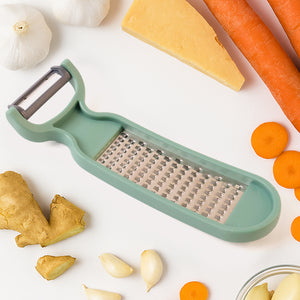 2-in-1 vegetable grater peeler in mint green with garlic, cheese, and carrots on a kitchen countertop
