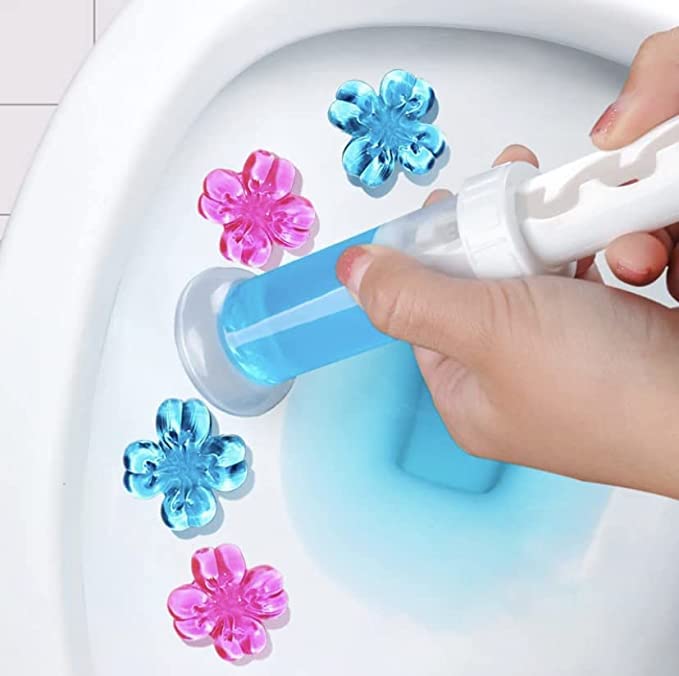 Blue and pink flower-shaped gel toilet stamps being applied to a white bowl with a hand-held plastic applicator.