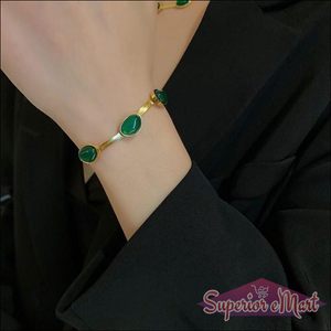 Gold-toned flat chain bracelet with three oval green stones worn on a wrist against a black blazer.