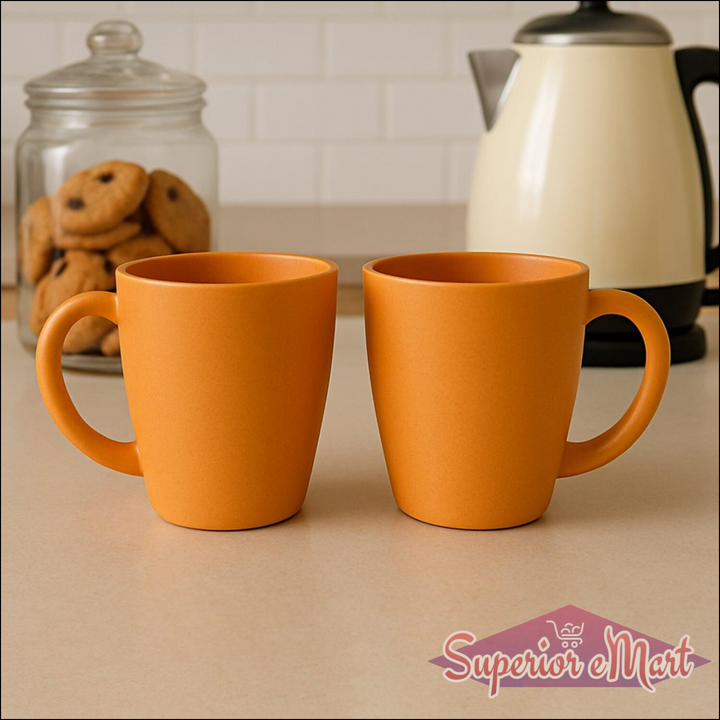 Set of two orange ceramic coffee and tea mugs set on a kitchen counter with a teapot and cookies
