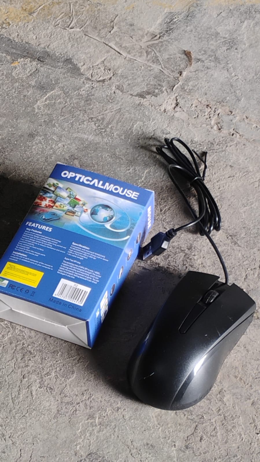 X2 computer mouse in black next to its blue packaging on a gray surface