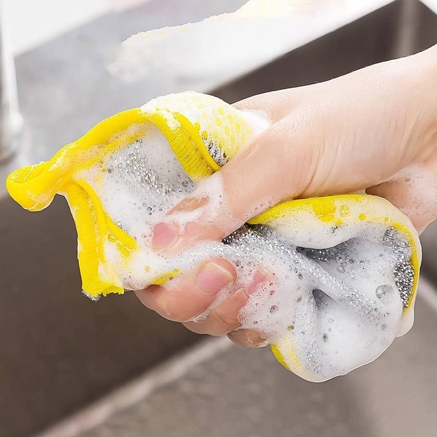 Hand squeezing a yellow and gray double-sided dishwashing sponge with soap suds over a kitchen sink