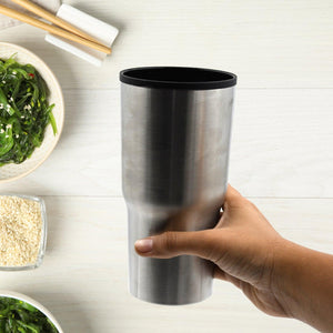 Hand holding a silver double walled travel coffee mug on a wooden table with bowls of greens