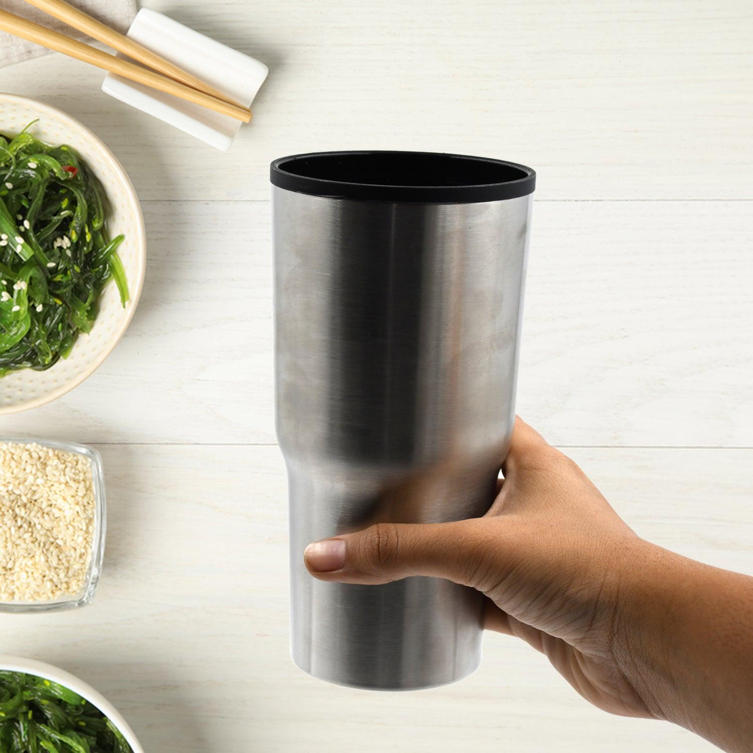 Hand holding a silver double walled travel coffee mug on a wooden table with bowls of greens