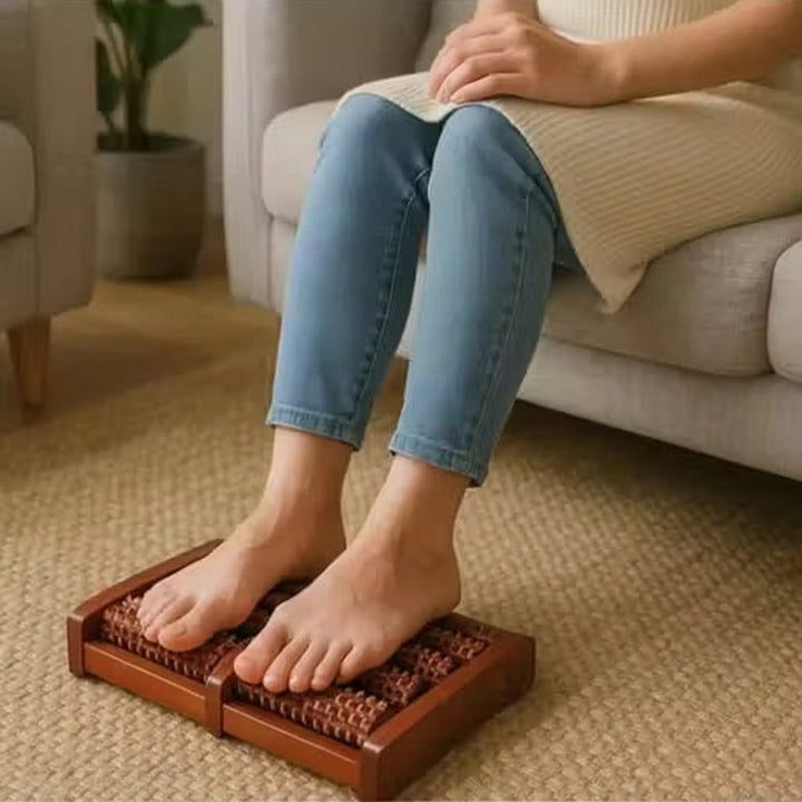 A person sitting on a sofa in a living room uses a wooden foot massager with textured rollers on their feet.