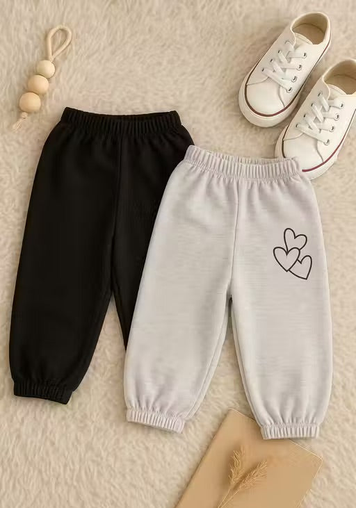 Two pairs of children's jogger pants laid flat on a soft cream background: one black pair and one light gray pair with a small heart outline graphic on the upper leg. White lace-up shoes, a wooden bead charm, and a small beige book or card are placed around them as styling props.