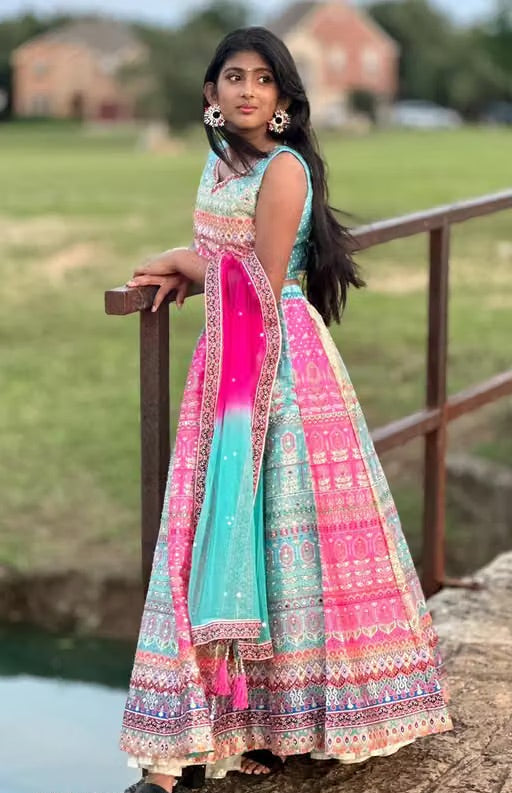 A woman wearing a vibrant pink, teal, and multicolor printed Anarkali gown with a matching dupatta, posing outdoors on a stone bridge with a grassy field in the background.