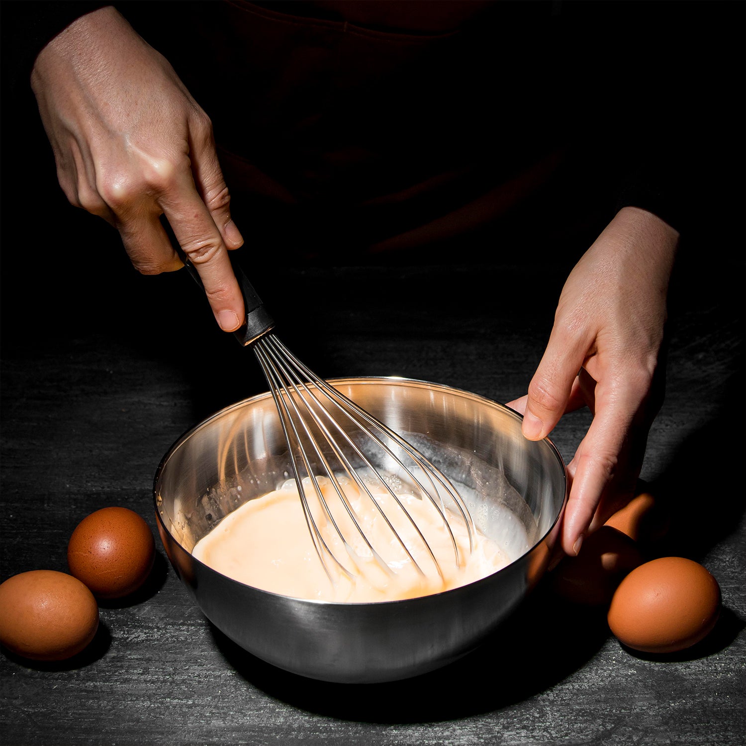 Stainless steel hand whisk being used to mix eggs in a bowl surrounded by eggs