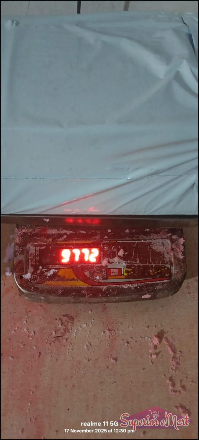 White poly mailer on a digital scale showing 9772 in red. Metal scale and floor with pink powder. Realme 11 5G watermark.