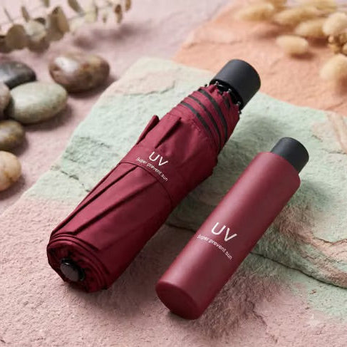 A folded dark red compact umbrella and a matching cylindrical UV sunscreen bottle with a black cap, styled on a textured pink surface with decorative stones and dried leaves.