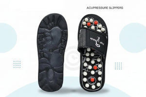 A pair of black acupressure slippers shown on a light blue and white promotional background. One slipper is displayed from the sole side, showing a textured footbed design, and the other is shown from the top side, revealing circular massage nodes in black, white, and red.