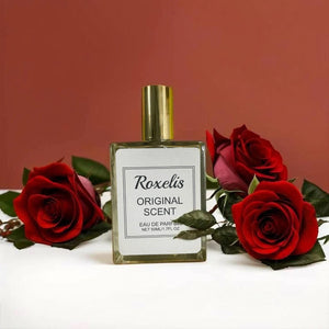 Roxelis Original perfume bottle surrounded by fresh red roses on a white surface