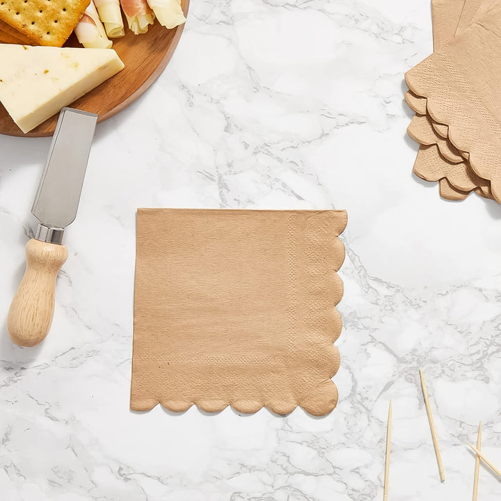 Scalloped edge disposable napkins in tan color on a marble table with a cheese platter