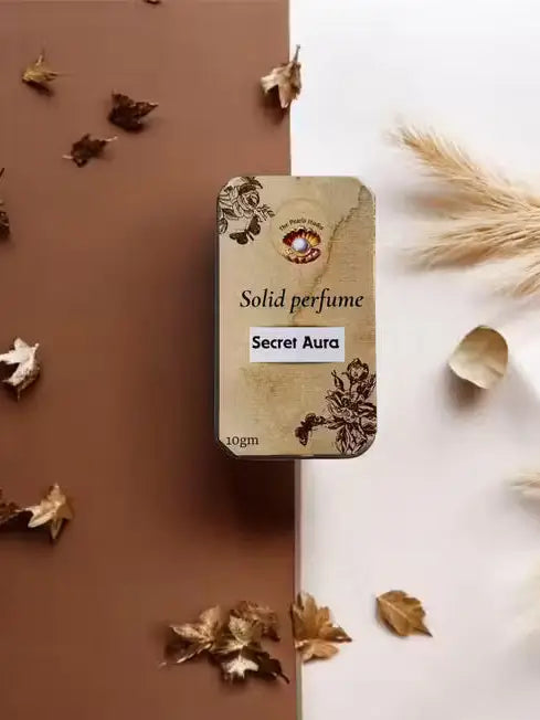 The Pearls Studio Secret Aura 10g solid perfume in a floral gold tin, surrounded by dried autumn leaves.