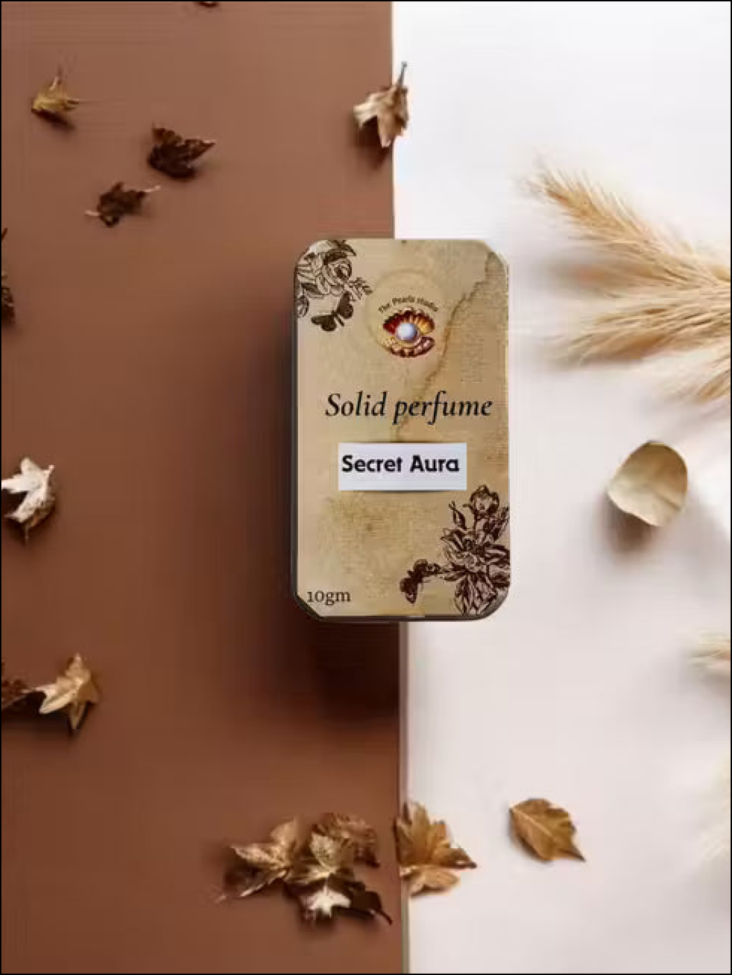 The Pearls Studio Secret Aura 10g solid perfume in a floral gold tin, surrounded by dried autumn leaves.