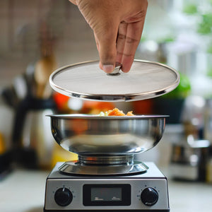Hand lifting a stainless steel mesh splatter screen over a stainless steel bowl on a stove