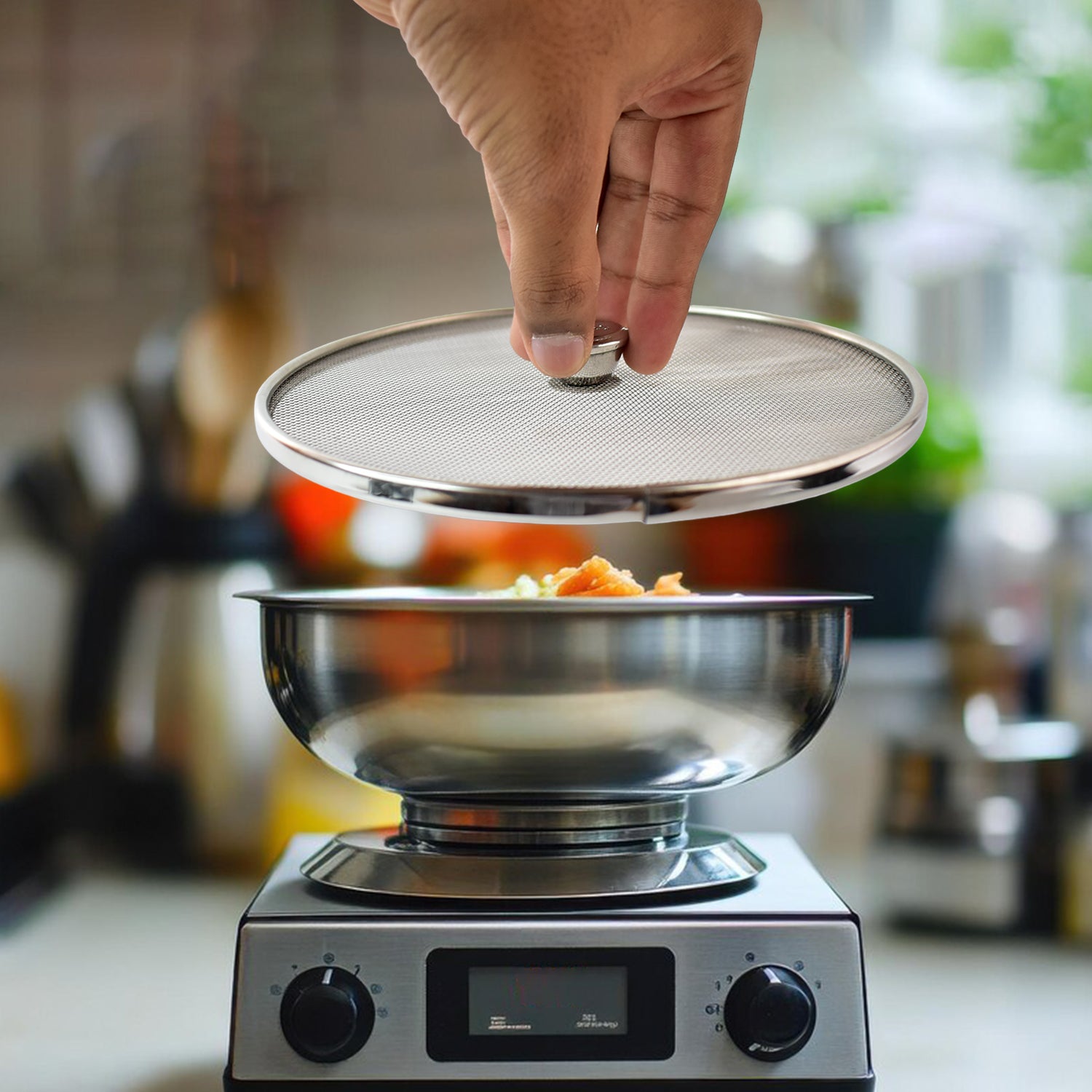 Hand lifting a stainless steel mesh splatter screen over a stainless steel bowl on a stove