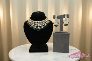 Ornate gold necklace and earring set with diamonds, green emerald beads, and red rubies on velvet display stands.