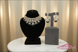 Ornate gold necklace and earring set with diamonds, green emerald beads, and red rubies on velvet display stands.