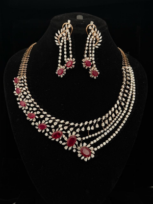 Gold jewelry set with diamond leaf motifs and red oval rubies, including an asymmetrical necklace and drop earrings.