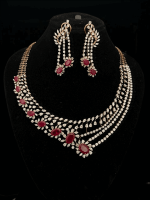 Gold jewelry set with diamond leaf motifs and red oval rubies, including an asymmetrical necklace and drop earrings.
