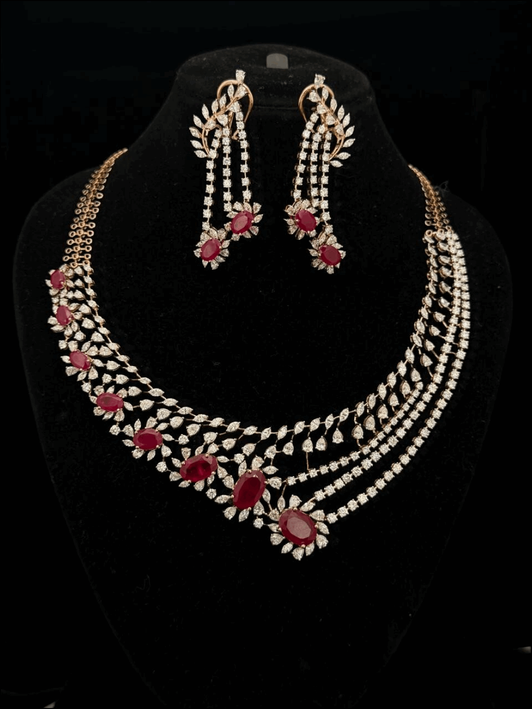 Gold jewelry set with diamond leaf motifs and red oval rubies, including an asymmetrical necklace and drop earrings.