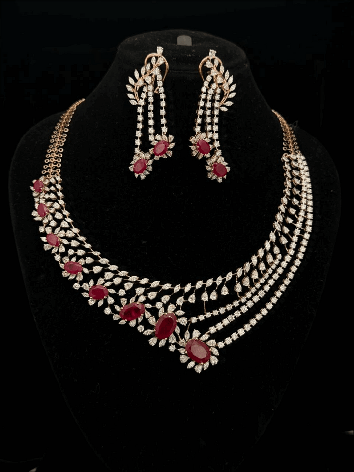 Gold jewelry set with diamond leaf motifs and red oval rubies, including an asymmetrical necklace and drop earrings.