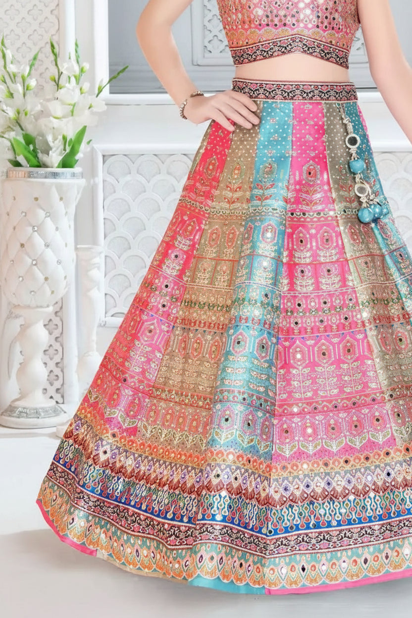 A close-up of a model wearing a vibrant pink, blue, and gold heavily embroidered Indian lehenga choli skirt set. The skirt features intricate mirror work and geometric patterns in pink, teal blue, and gold, with a matching embroidered top partially visible.