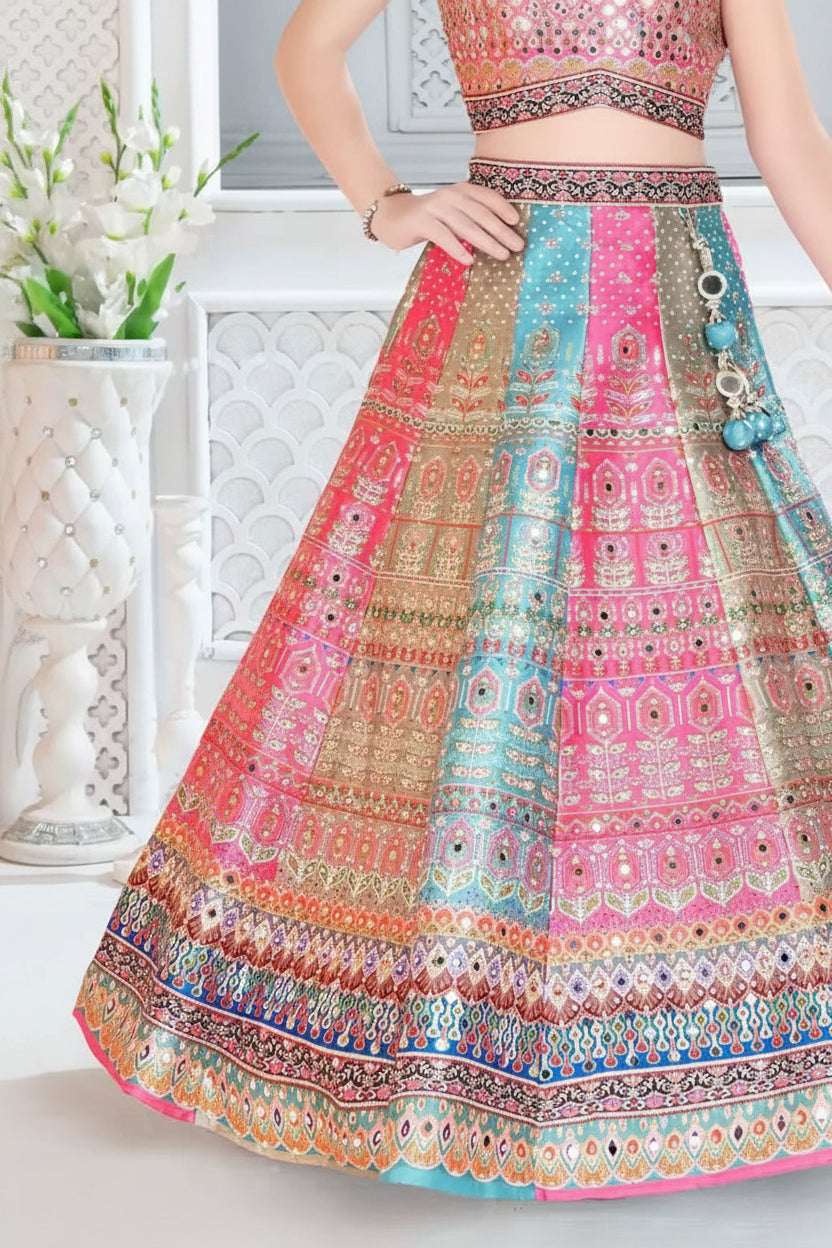 A close-up of a model wearing a vibrant pink, blue, and gold heavily embroidered Indian lehenga choli skirt set. The skirt features intricate mirror work and geometric patterns in pink, teal blue, and gold, with a matching embroidered top partially visible.